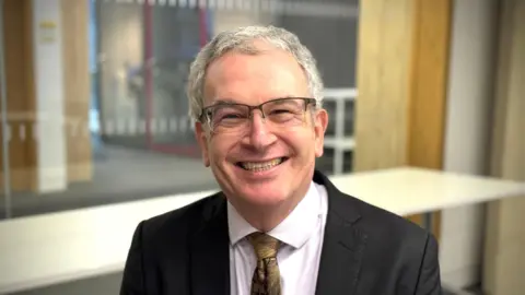 Prof David Reiner is sitting in a boardroom at Judge Business School where he is professor of technology policy. He is wearing a black suit and glasses and is smiling