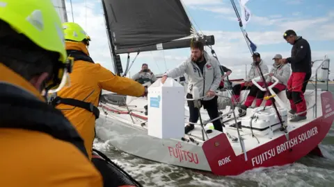 Two lifeboat crew members are shown left wearing yellow safety gear facing away from the camera as one reaches for a white box being passed to him by a member of a yacht alongside them at sea. The yacht has red and white livery with a grey sail. Four male crew members can be seen sitting behind the man.