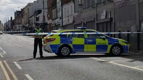 Police tape strung across Shields Road in Byker. A police officer stands next to a marked police car behind the cordon.