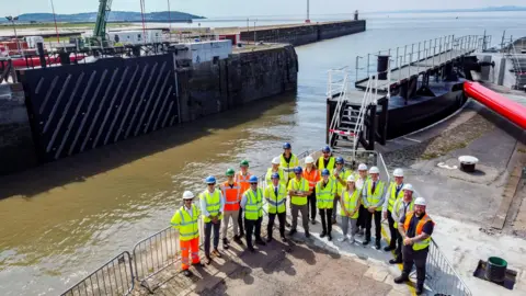 A group of workers in high-vis jackets are seen standing by the new dock gates at Avonmouth Port. The Severn Estuary is visible in the background