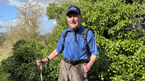 Mike Patrick on a sunny day outdoors in nature with greenery in the background. He is wearing a blue polo shirt, a Cure EB cap and is carrying walking poles. 