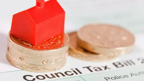 A red Monopoly house sits on top of three pounds coins, next to two pound coins, all placed on top of a council tax bill.