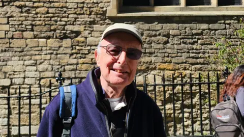 Eoin McCaul/LDRS A man, wearing a beige hat and sunglasses, stares at the camera. He wears a navy fleece and has a strap of a backpack on one shoulder. He stands in front of a historic brick wall and a black railing.