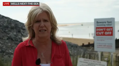 BBC reporter Debbie Tubby, who has shoulder-length blonde hair and is wearing a red jacket over a white top, reports live from a vantage point at the beach. A white and red sign is behind her, warning beach-goers of the dangers of the tide. Sand and water is even further in the background.