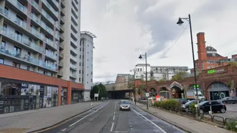 Google A car drives along the road, with high rise buildings at either side.