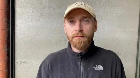 Dan Jennings has a beard and is looking directly into the camera. He wearing a zip up black fleece top and a beige baseball cap. He is stood against a concrete and red brick wall.