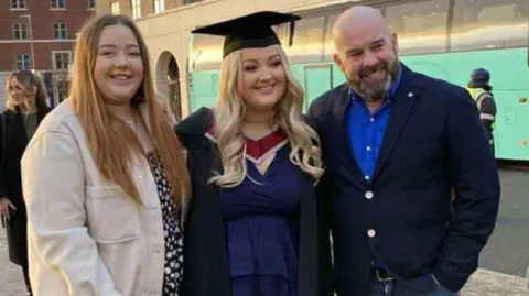 Family photo A girl with curly blonde hair wearing a blue dress and a graduation gown and cap. One side of her is a girl with long brown hair wearing a black dress and cream jacket, and on the other is a bald man with a grey beard, wearing a blue shirt, navy cardigan and blue jeans. They are all smiling, with their arms around each other.