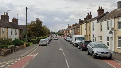 A residential street with a row of terraced houses on either side. In front are cars parked up on the side.