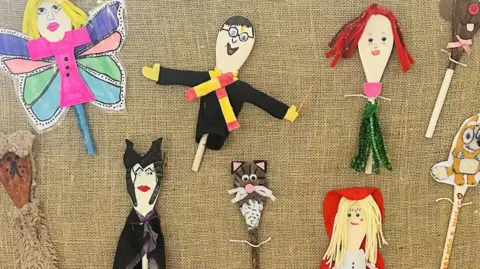 Bardwell Primary School Wooden spoons decorated to look like children's book characters lay on a flat surface covered in a brown sheet. The likes of Harry Potter, a cat, Maleficent, and more characters can be seen.
