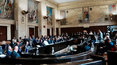 John Fairhall/BBC The Essex County Council chamber with paintings and old maps on the wall. Around 80 people are seated in a crescent around the top table