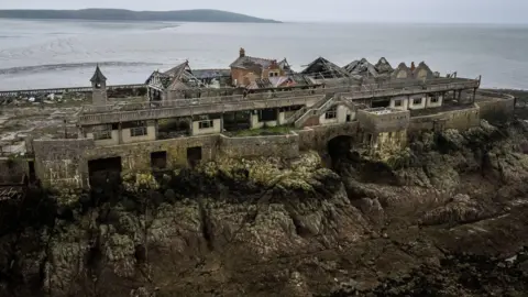 Press Association Buildings perched on the rocky island, set on large stone foundations. The buildings are in a severe state of decay, with broken roofs and empty windows. The sky and sea are grey. 