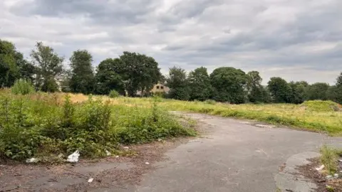 Phil Towers/BBC An unused piece of land, which has a small section that is concreted over. The rest is grass and weeds, and in the distance are trees which surround the site.