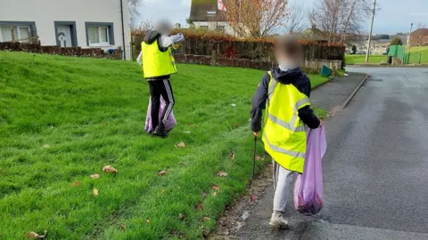 Two teenage boys wearing sports tracksuits and fluorescent jackets pick up litter in a residential street. They are holding purple bin bags which are becoming fuller with litter. 