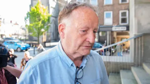 Pacemaker Anthony McGinn walking up concrete steps into a courtroom. The camera is close to his face as he looks away. He has messy brown and white hair. He is wearing a light blue short sleeve shirt and has a pair of black-rimmed glasses tucked into the front of his shirt.