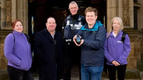 Two women and three men stand outside a town hall building as one of the men shows off a body-worn camera. The women wear purple fleeces and the men are in coats, one is a traffic warden.