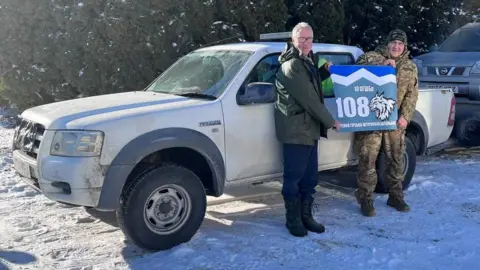 Bruce Burrow Bruce and a soldier stand in front of a car in the snow. They are both holding on to two corners of a blue sign which is written in Ukrainian.