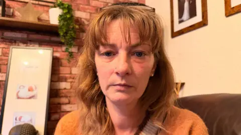 Head and shoulders shot of Helen. She is wearing an orange jumper and is looking directly at the camera. She is sitting on a brown-coloured sofa.