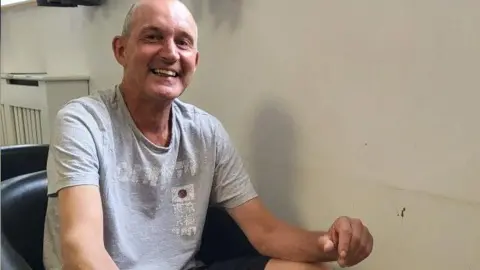 BBC Paul is sat down playing Connect Four, he is smiling and wearing a grey t-shirt and black shorts.