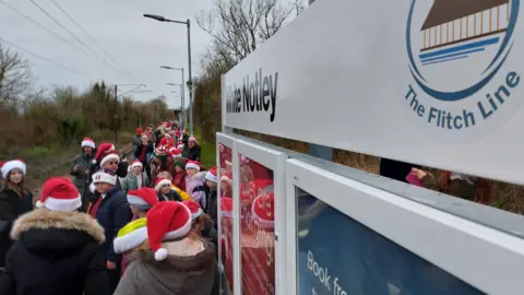 Scott Dolling Schoolchildren wearing Santa hats stood at the White Notley train station. In the foreground is a white sign, reading White Notley, and a crest reading "The Flitch Line"