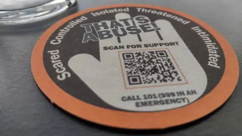 A round beer mat on a grey table. It is coloured black, white and orange and features a white illustration of a hand, along with a QR code and the messages: "That's Abuse!"; "Scared, controlled, isolated, threatened, intimidated"; "Scan for support"; and "Call 101 (999 in an emergency)". 