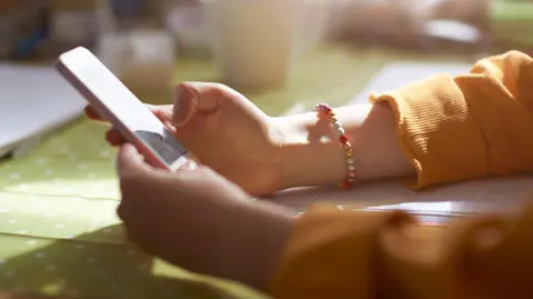 A young woman with a bangle round her arm in a yellow jumper is looking at a mobile phone