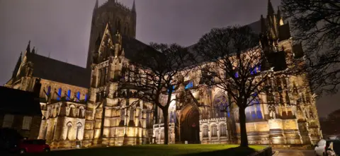 Helen Earth Lincoln Cathedral floodlit at night with two trees in silhouette. The cathedral is a large building built from honey coloured stone and has arched windows and columns. The tower is reaching into the dark night sky. 