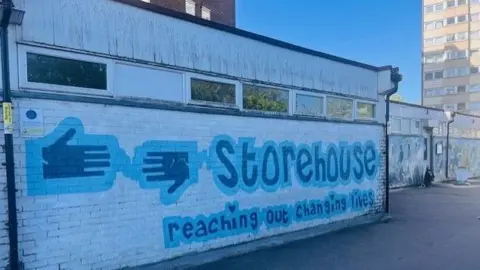 The Southend Vineyard Storehouse A predominately white building, with "Storehouse" and reaching out changing lives" painted in blue on the front. Two hands are also painted in blue on the front. There is a high rise building ion the background. The sky is blue.