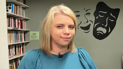 A young woman with short blonde hair wearing a turquoise dress standing in front of a green wall featuring painted theatre faces 