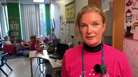  Ros Hawley faces the camera, she wears a pink t-shirt. Her blonde hair is pulled back from her face.