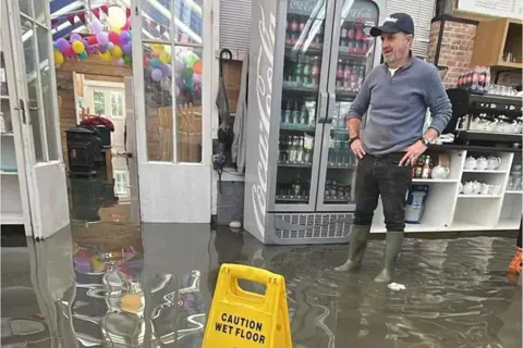 A man wearing wellies stands with his hands on his hips looking across a flooded coffee shop. The water is shallow but it is brown and covering the entire floor.