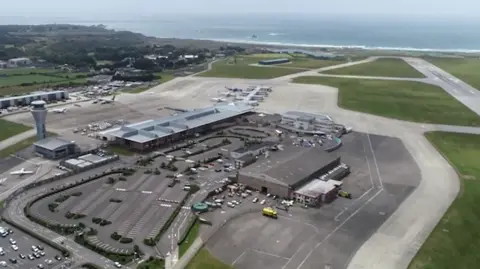 An aerial view of Jersey's airport surrounded by green fields on the right and fields and buildings on the left, with the ocean in the distance.