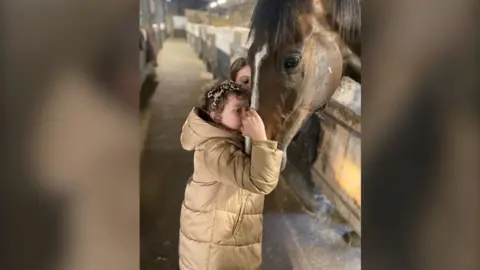 Young girl Betsy, wearing a puffer coat and holding her racehorse's face close to her. 