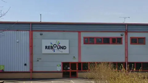 Google The outside of the Rebound Trampoline Park building. It is an industrial building with red windows. It has a sign on it that reads "REBOUND". The sky is blue.