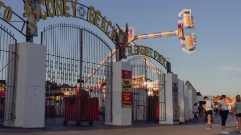 The gates to Coney Beach Amusement Park in the late afternoon sun. People are socialising in front of the entrance to a fairground with a revolving gondola ride spinning against a clear blue sky.