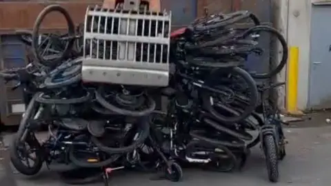 Multiple confiscated e-bikes being crushed. A pile of bikes can be seen with a digger bucket on top of them. 