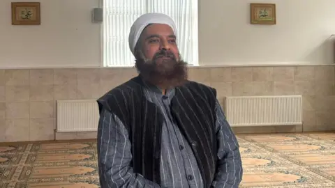Afaq Khan An imam waring a white turban and jacket sits inside a mosque. 