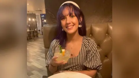 A young girl sitting in a restaurant in a brown leather booth smiling at the camera and holding a large glass with a piece of lemon in it. She has purple hair with a white headband and white dangly earrings and is wearing a black and white chequered top.