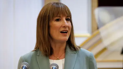 Chancellor Rachel Reeves wearing a sage green jacket and white collarless blouse stands behind a podium with two microphones giving a speech at the annual Mansion House dinner in the City of London, UK.
