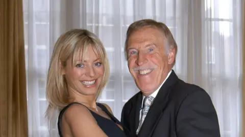 Daly smiles at the camera while posing in a dance position alongside Bruce Forsyth. She has short blonde hair and wears a black dress. Forsyth has short grey hair and wears a black suit and tie.