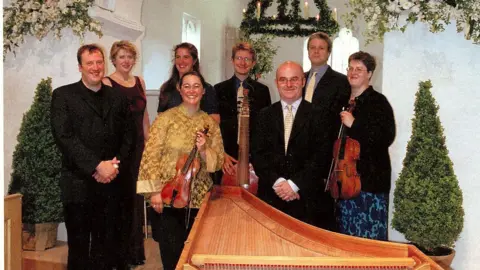 Maggie Faultless People dressed smartly and holding instruments are pictured standing in a what looks like a church building with whitewashed walls. There are potted trees and white and green hanging flowers surrounding them. All of the people in the image are smiling.