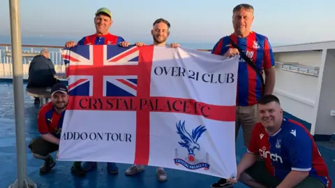 Harry Green Five people on an outdoor deck with a sea view hold up a large Crystal Palace FC flag with the words “OVER 21 CLUB” and “ADDO ON TOUR,” while others crouch beside it. All are wearing red-and-blue football shirts.