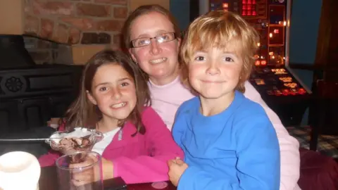 Karl Lansley Carli Lansley in the centre of frame, with her daughter on the left and her son on the right, sitting at a table in a pub or restaurant. There is a brick fireplace and a gambling machine behind them. Carli is wearing a pink jumper and has dark-rimmed rectangular glasses, and has brown shoulder-length hair. Her daughter has dark brown shoulder-length hair, is wearing a white T-shirt with pink cardigan on top, and is holding an ice cream dessert. Carli's son is wearing a light blue round-neck jumper and has touseled blonde hair. All three are smiling at the camera.