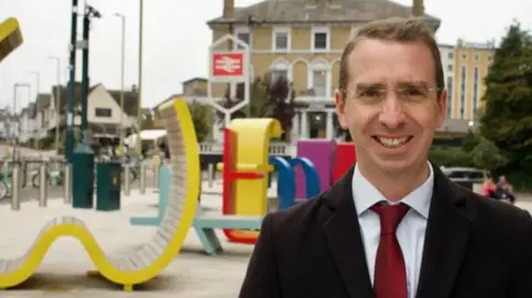 Peter Taylor with short dark hair smiling at the camera while wearing a dark jacket, white shirt and red tie. His is standing in front of a railway station and some yellow plastic shapes installed as play equipment in the street.