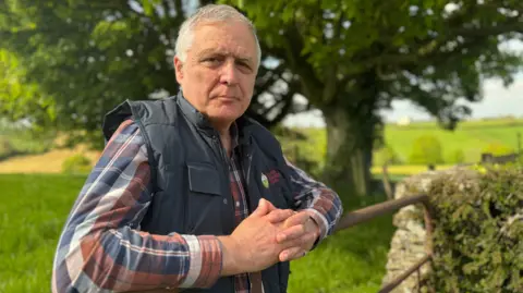 William is standing in a grassy field and is leaning over a metal gate. He is wearing a colourful plaid shirt and navy gilet. He has grey hair. 