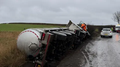 Sebastian Cruddas A HGV tanker overturned on a country road. The vehicle has fallen on its side into a ditch and has flattened vegetation. Other vehicles can be seen further along the road. A person in an orange hi-vis top and bottoms is perching on a front wheel looking into the cab. 