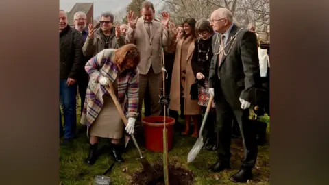 A group of people plant a tree including, on the right, a man in a suit and mayoral chains