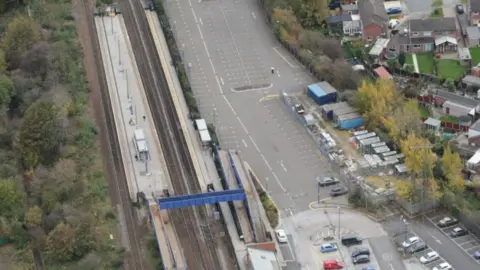 Ariel view of Swinton railway station in Rotherham. A blue footbridge is crossing the tracks between platforms. The station car park is also in view, with a majority of the parking spaces empty. Nearby houses can also be seen. On the other side of the railway line are trees