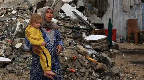 Reuters A Palestinian woman holds a child dressed in a yellow track suit, with a pile of rubble behind them.