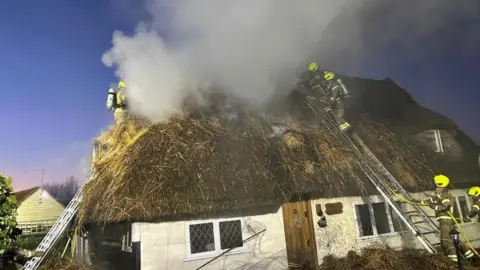 A white cottage with a thatched roof which is covered in smoke. Three firefighters are on the roof and two further ones are on the ground. 