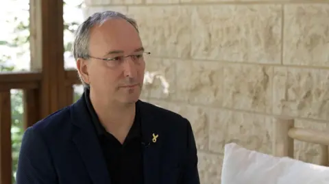 Eran Etzion wears a dark navy blue suit and polo shirt as he speaks to the BBC